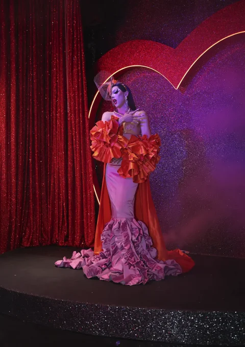 Drag performer in a pink-and-orange gown with large ruffles posing on a glittery stage with red sequin curtains and a neon arch backdrop from the audience view of a show